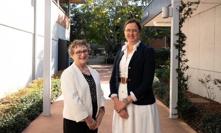 Two women stand outdoors in a shaded walkway between buildings, both dressed in professional attire and smiling at the camera.