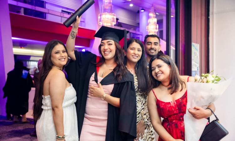 Five people pose indoors; one in graduation cap and gown holds a diploma, while another holds a bouquet of flowers. All are smiling at the camera.