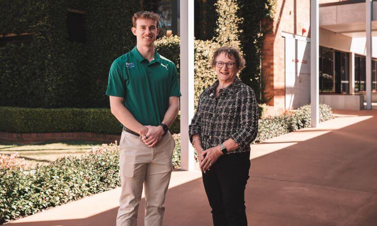 Two people, one wearing a green polo shirt and khaki pants and the other in a patterned blouse and black pants, stand outside on a pathway near a building and greenery.