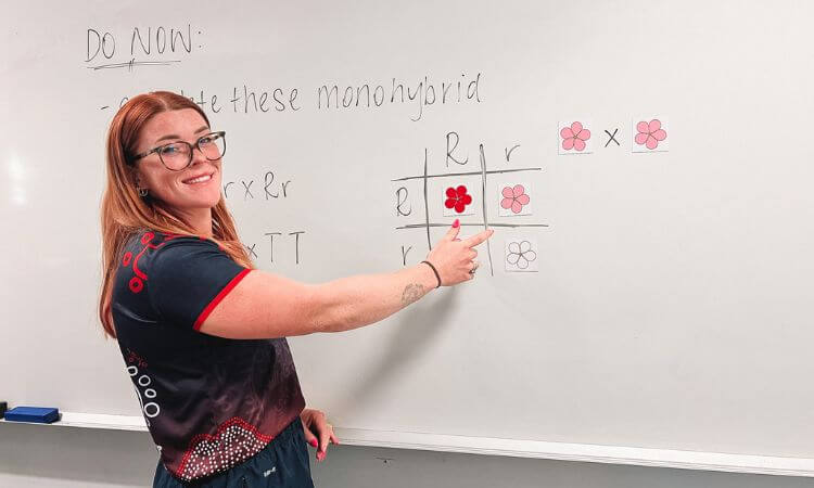A woman standing at a whiteboard points to a Punnett square diagram illustrating monohybrid genetic crosses with flower traits.
