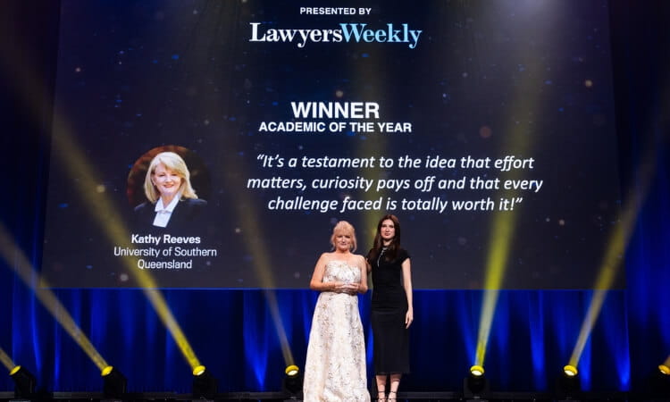 Two women stand on stage at a Lawyers Weekly event, presenting the Academic of the Year award to Kathy Reeves from the University of Southern Queensland. Her photo and quote are displayed on a screen.
