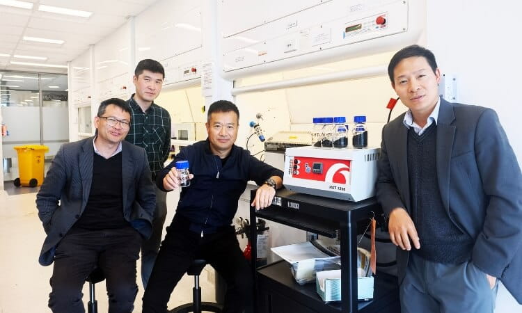 Four men pose in a laboratory with scientific equipment and sample bottles on a cart; lab benches and supplies are visible in the background.
