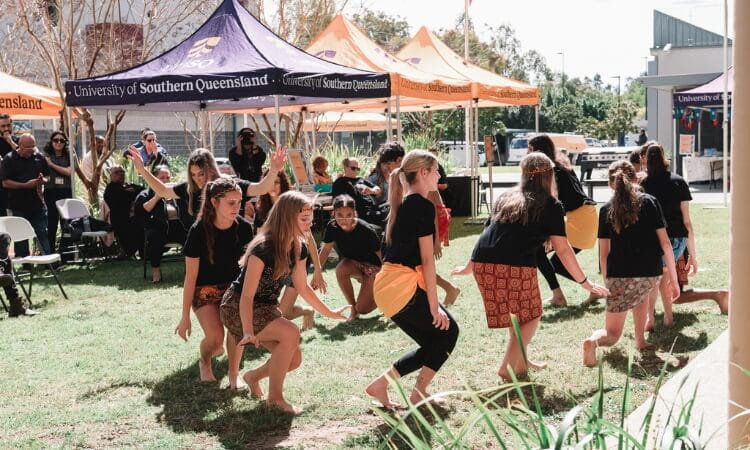 A group of young people perform a dance outdoors in front of University of Southern Queensland tents, with spectators seated in the background.