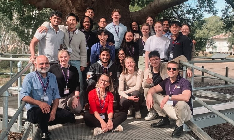 A group of twenty-three people pose together outdoors on a metal ramp in front of a large tree, all wearing name badges.