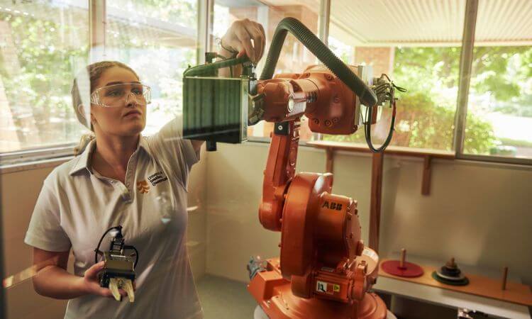 A woman wearing safety glasses operates an industrial robotic arm inside a laboratory with large windows.
