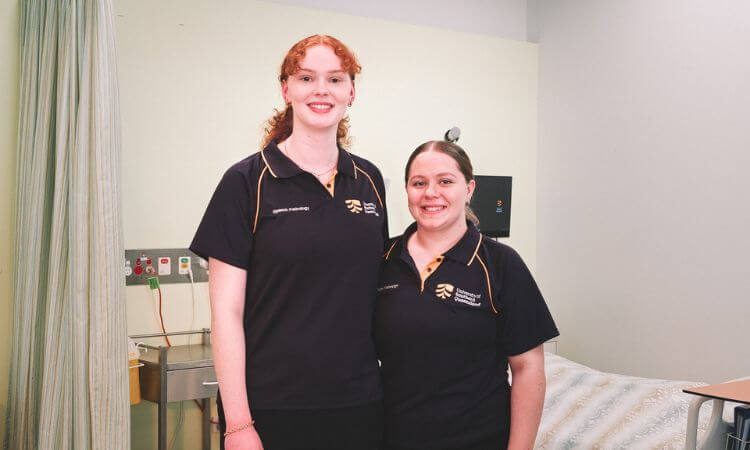 Two women in matching navy uniforms stand side by side in a clinical room with a hospital bed and medical equipment in the background.