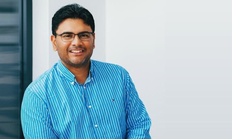 A man wearing glasses and a blue striped shirt smiles while standing in front of a plain light-colored background.