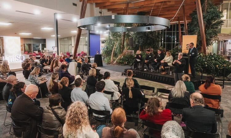 A group of people listen to a panel discussion in a modern indoor venue with rows of chairs and plants in the background.