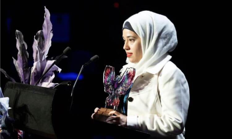 A woman in a white coat and headscarf stands at a podium, holding a colorful eagle-shaped award, speaking into a microphone.
