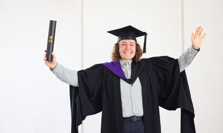 Person in graduation cap and gown holds a testamur in one hand and raises the other arm, smiling against a plain white background.