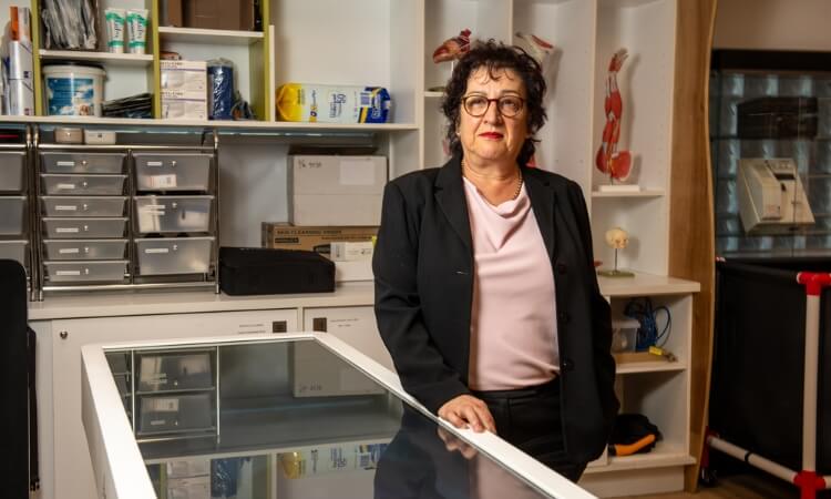 A woman wearing glasses and business attire stands in a medical office, with shelves containing supplies and anatomical models in the background.