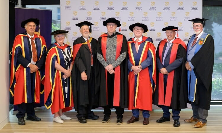 Seven people in academic regalia and hats stand in a row indoors, posing for a group photo against a backdrop with a university logo.