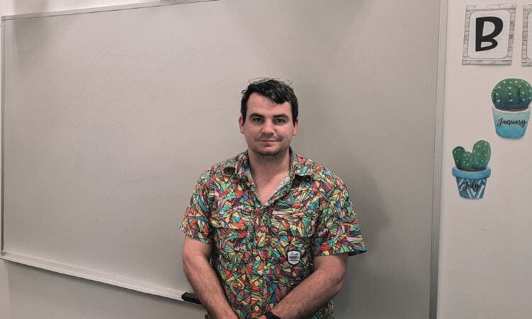 A man in a colorful shirt stands in front of a whiteboard, with wall decorations including cactus drawings and a letter "B" visible on the right.