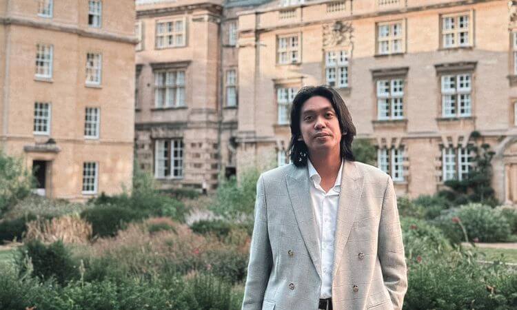 A person in a light gray suit stands in a garden with an old, beige stone building in the background.