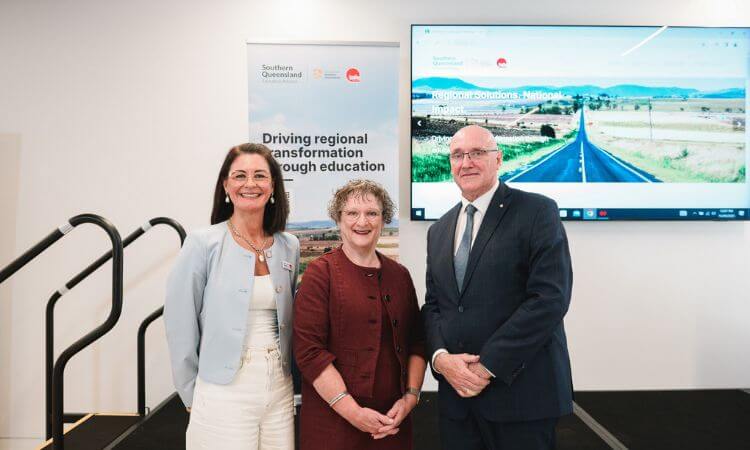 Three people in business attire stand indoors in front of a promotional banner and a screen displaying a rural landscape, at an education-focused event.