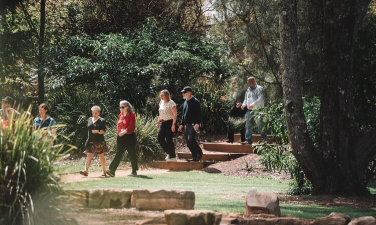 A group of people walk along a stone path through a garden with trees and greenery during daylight.