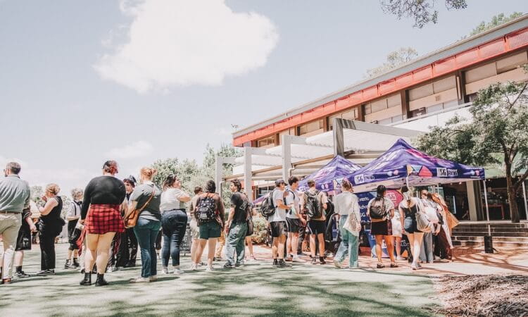 A line of people stands outdoors near two purple canopy tents in front of a building on a sunny day.