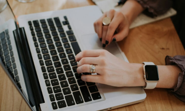 Person wearing a smartwatch and rings types on a laptop keyboard with one hand while writing in a notebook with the other.