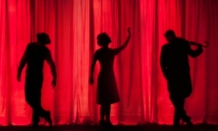 Three people in silhouette pose dramatically in front of a red stage curtain, with one in the center raising an arm and the other two leaning to either side.