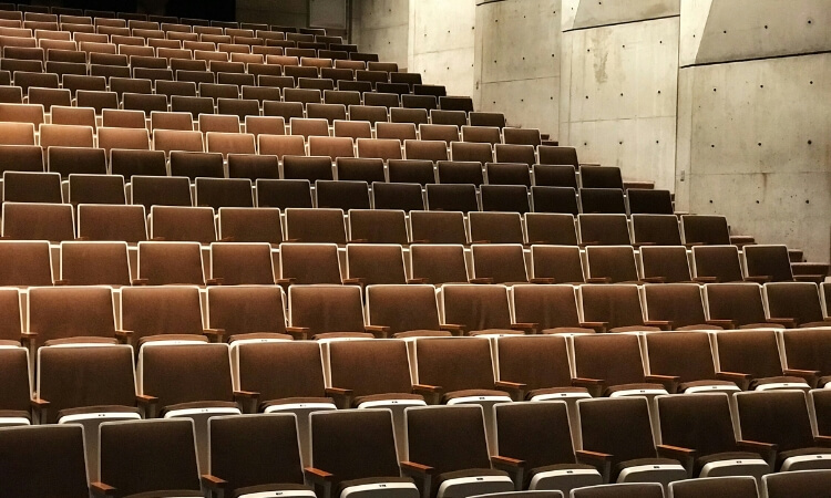 Rows of empty brown auditorium seats arranged in tiers, with a concrete wall on the right side.