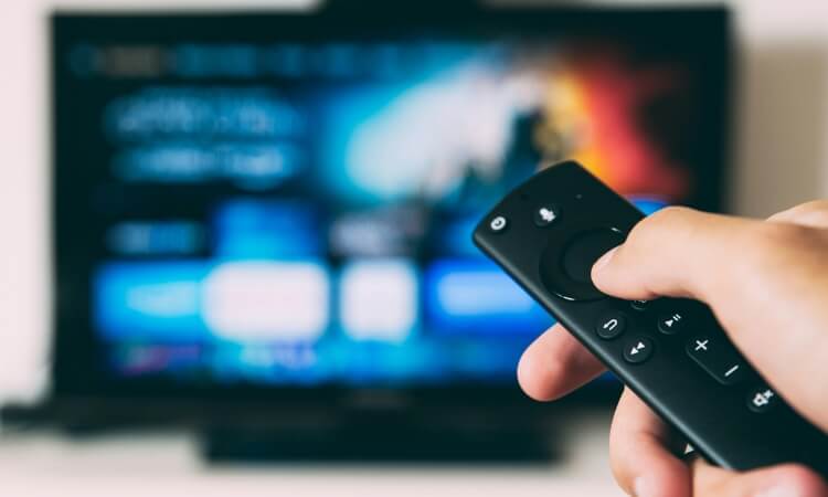 A hand holding a TV remote control, pointed at a television screen displaying a blurred streaming service menu.