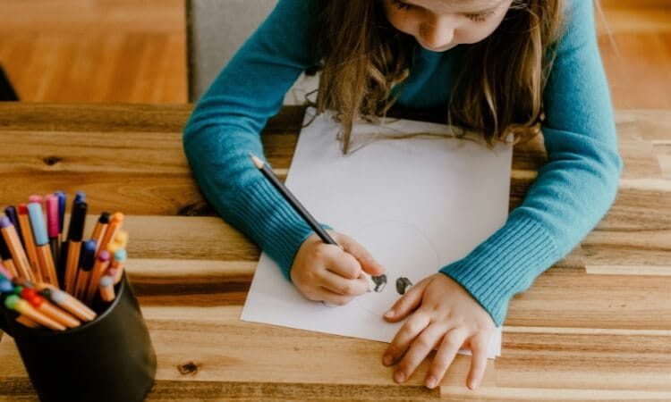 Young child in a blue sweater sits at a wooden table, drawing on white paper with a pencil; a cup of colored markers is nearby.