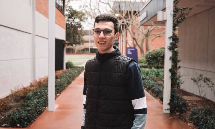 A young man wearing glasses and a black vest stands outside on a walkway between buildings, with trees and shrubs in the background.