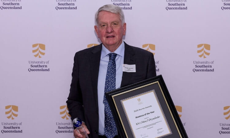 An older man in a suit holds an award and a framed certificate in front of a University of Southern Queensland backdrop.