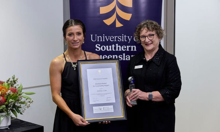 Two women stand indoors; one holds a framed award certificate and the other holds a trophy. A University of Southern Queensland banner is visible in the background.
