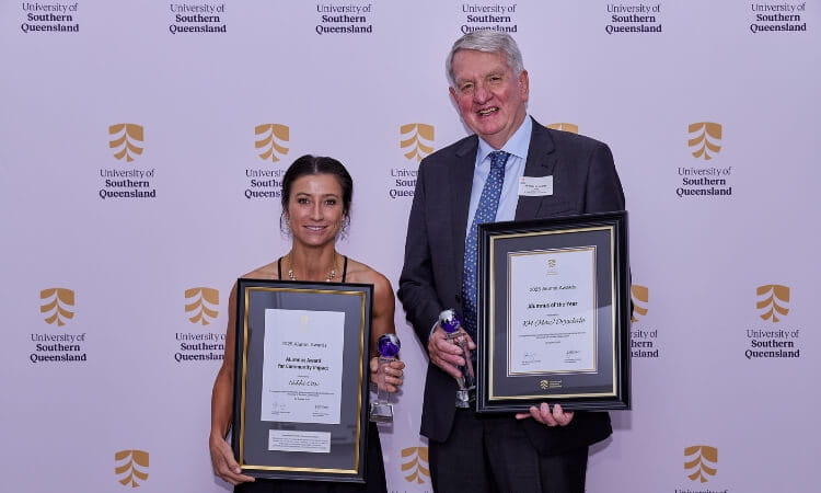 Two people stand in front of a University of Southern Queensland backdrop, holding framed awards and glass trophies, smiling at the camera.
