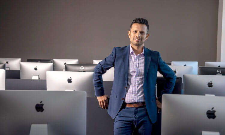 A man in a suit stands in a computer lab with several iMac computers, facing the camera and leaning on one of the desks.