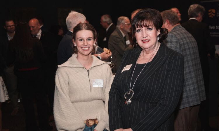 Two women pose for a photo at a formal event with a group of people conversing in the background.