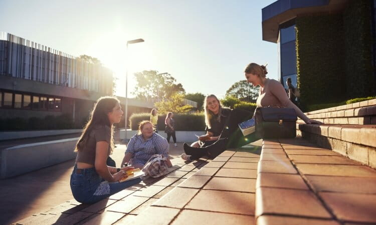 Four people sit and talk on outdoor steps on a sunny day, with books and bags around them, near modern campus buildings.