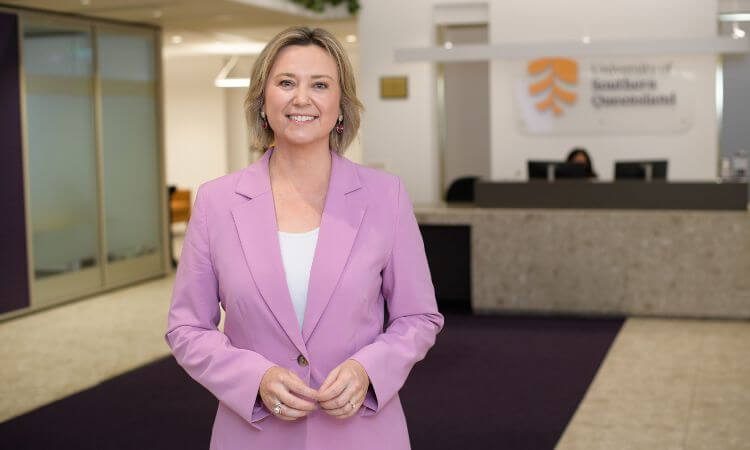 A woman in a pink blazer stands and smiles in an office lobby with a University of Southern Queensland sign in the background.