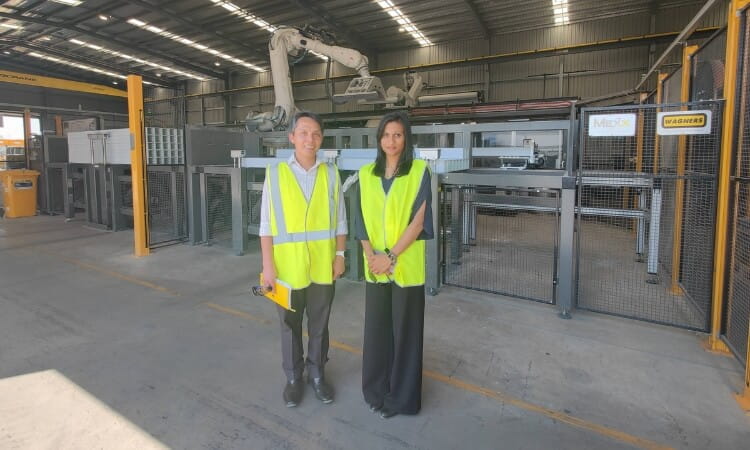 Two people in yellow safety vests stand in front of industrial machinery inside a large factory or warehouse.