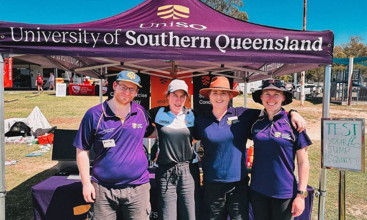 Four people stand in front of a University of Southern Queensland tent, smiling at the camera, with an outdoor event setup in the background.