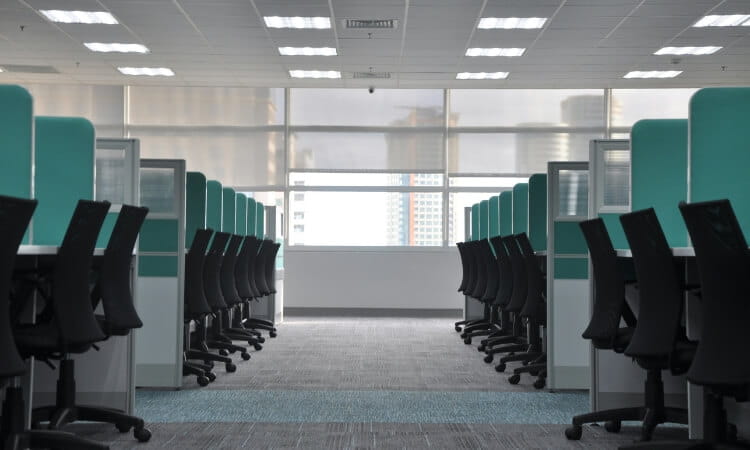 Empty office with rows of black chairs and desks separated by teal dividers, illuminated by ceiling lights, and a large window at the far end.