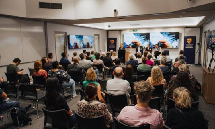 Audience seated in a conference room facing a panel of speakers at the front, with presentation slides displayed on screens behind them.