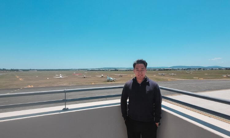 A man stands on a balcony overlooking an airfield with several small planes on the runway under a clear blue sky.