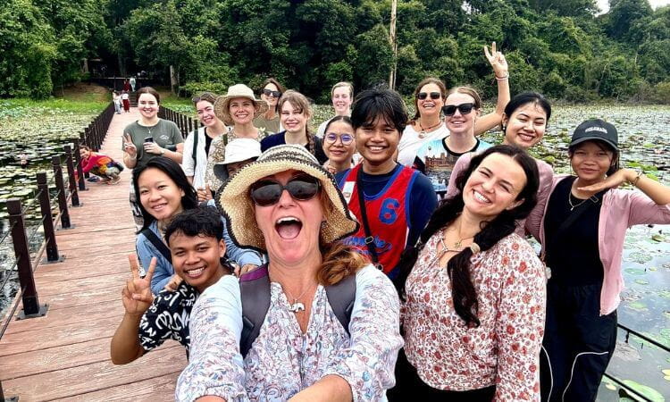 A group of people smiling and posing for a selfie on a wooden bridge over a pond with lily pads, surrounded by lush green trees.