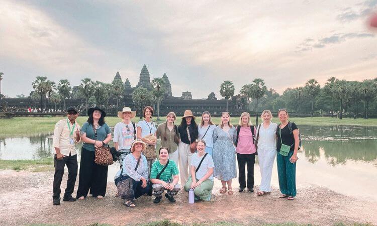A group of people poses for a photo in front of Angkor Wat, with palm trees and a pond in the background under a cloudy sky.