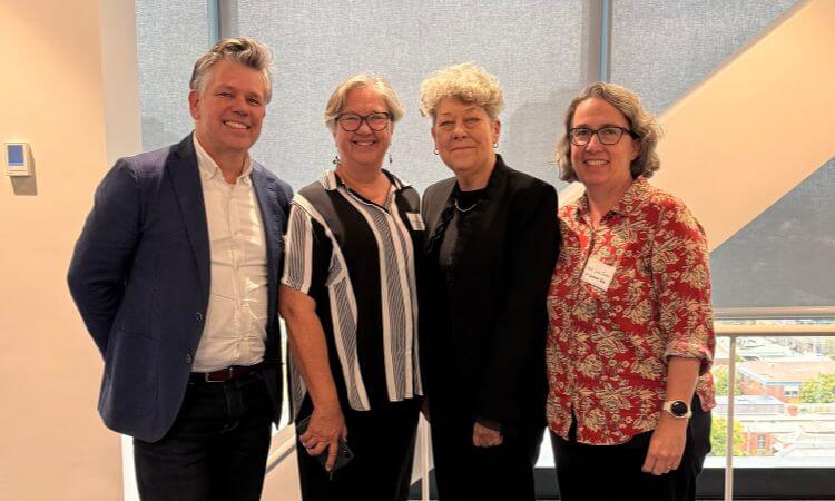 Four adults stand indoors, smiling for a group photo in front of a large window with city buildings visible outside.