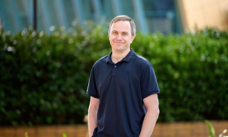 A man wearing a dark polo shirt stands outdoors in front of green bushes and blurred modern buildings, smiling at the camera.