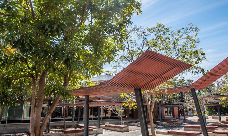 Outdoor courtyard with angled metal shade structures, large trees, and a building with glass windows in the background under a blue sky.
