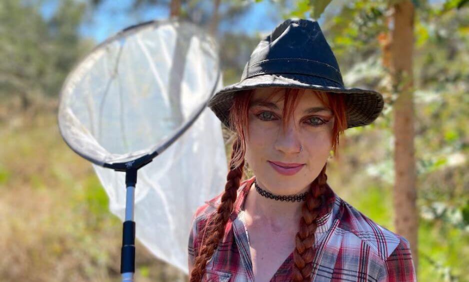 Person wearing a black hat and plaid shirt with braided hair stands outdoors holding a butterfly net over their shoulder. Trees and foliage are visible in the background.