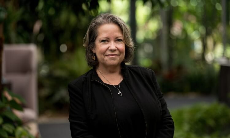 A woman with shoulder-length brown hair wearing a black jacket stands outdoors, smiling, with greenery and blurred plants in the background.