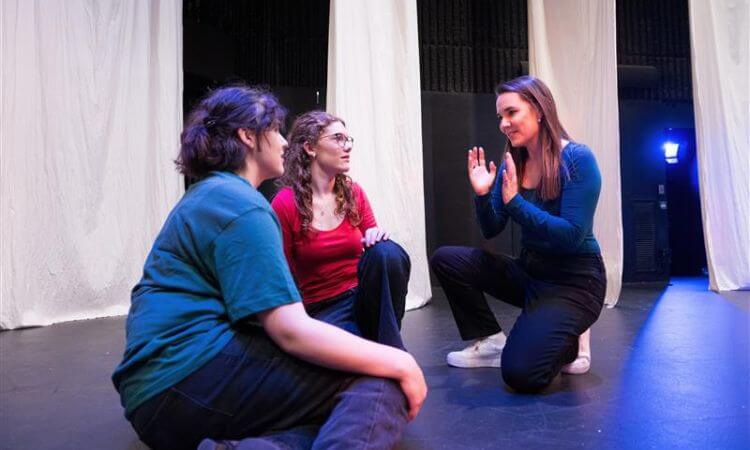 Three women sit and kneel on a stage with white curtains in the background, engaged in a focused conversation or rehearsal.