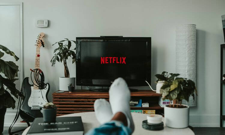 Person with feet up on a table watching Netflix on a TV in a living room with plants, a guitar, and modern decor.