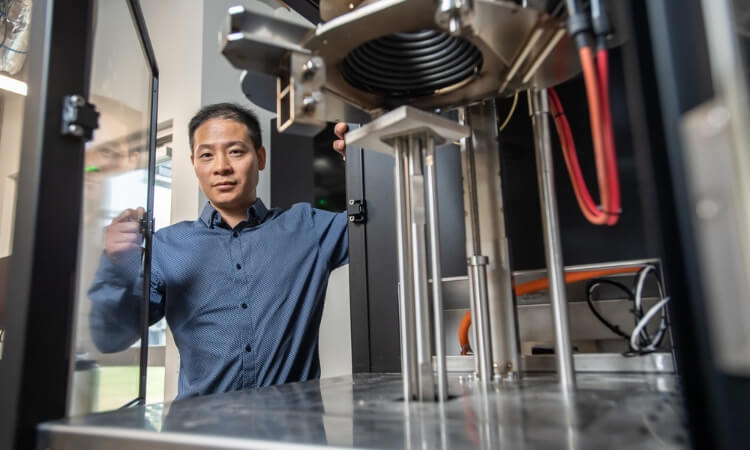 A man in a blue shirt stands next to industrial equipment, looking at the camera in a laboratory or manufacturing setting.