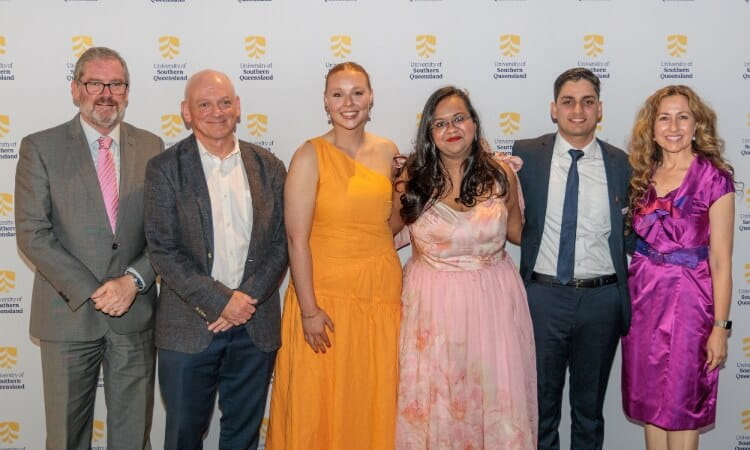 Six people stand in a row, dressed in formal attire, smiling for a group photo in front of a University of Southern Queensland backdrop.